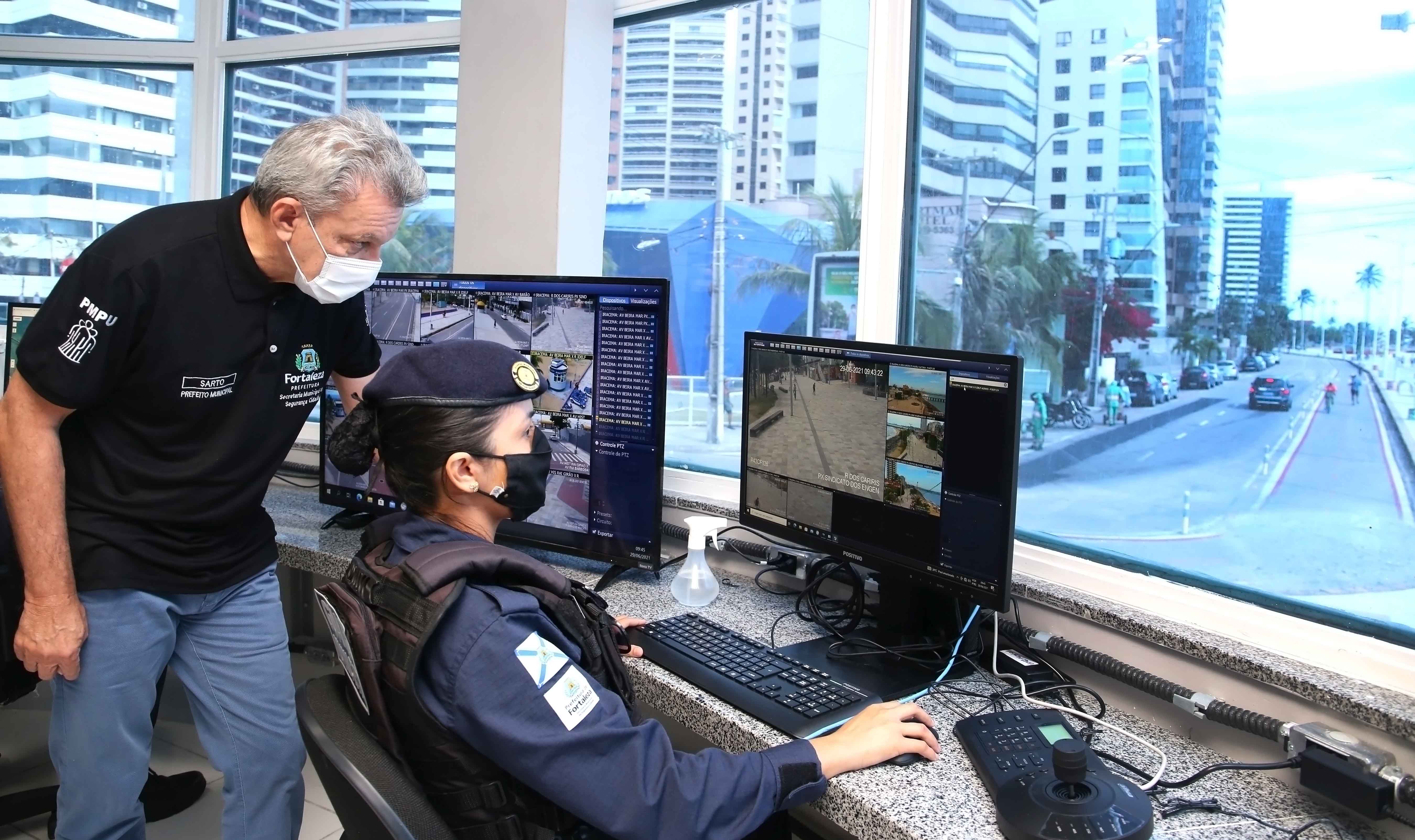prefeito Sarto observa uma tela de videomonitoramento dentro de uma célula de segurança. Uma guarda municipal na frente da tela explica para ele o funcionamento do equipamento 